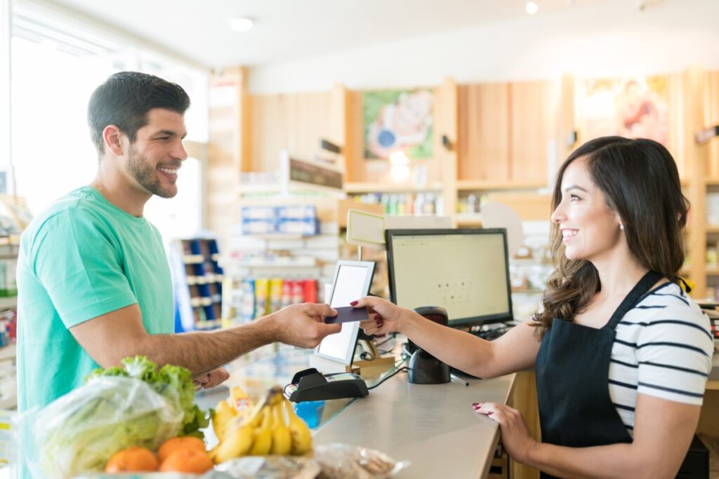 A customer in a green shirt hands a credit card to a cashier wearing an apron and striped shirt at a grocery store checkout counter. Various groceries, including bananas, lettuce, and oranges, are on the counter. Shelves with products and bright lighting are visible in the background.