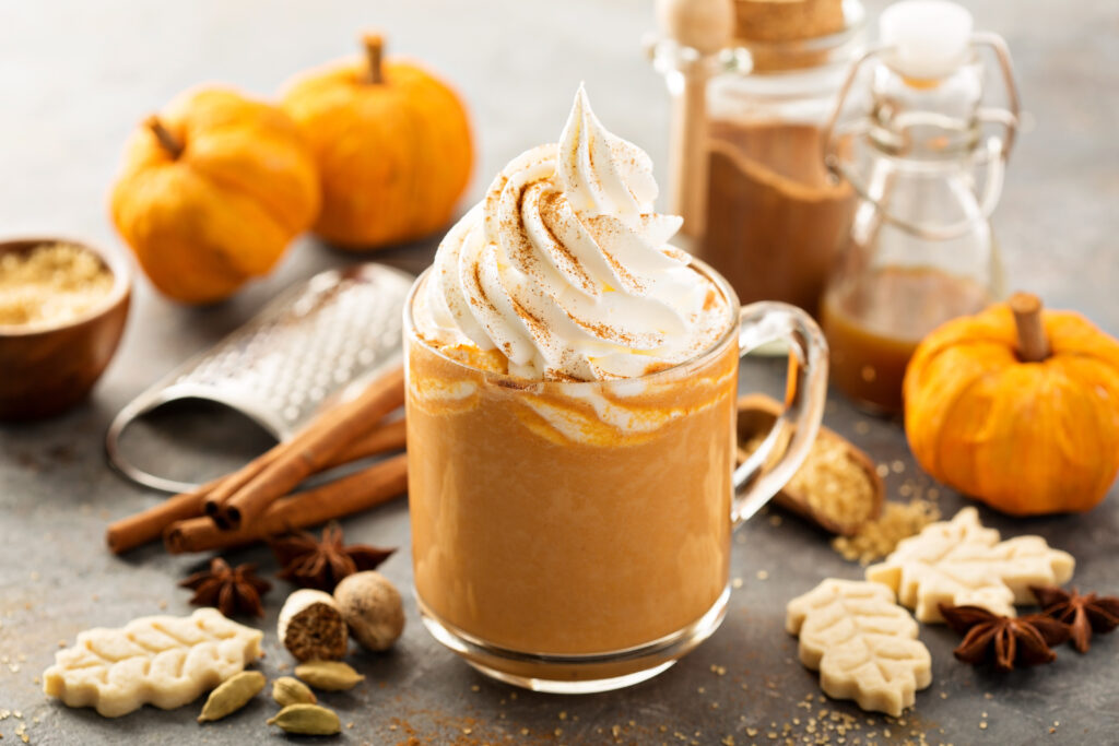 A cozy autumn scene featuring a glass mug filled with pumpkin spice latte topped with whipped cream and sprinkled cinnamon, surrounded by pumpkins, cinnamon sticks, nutmeg, star anise, brown sugar, and fall-themed cookies.