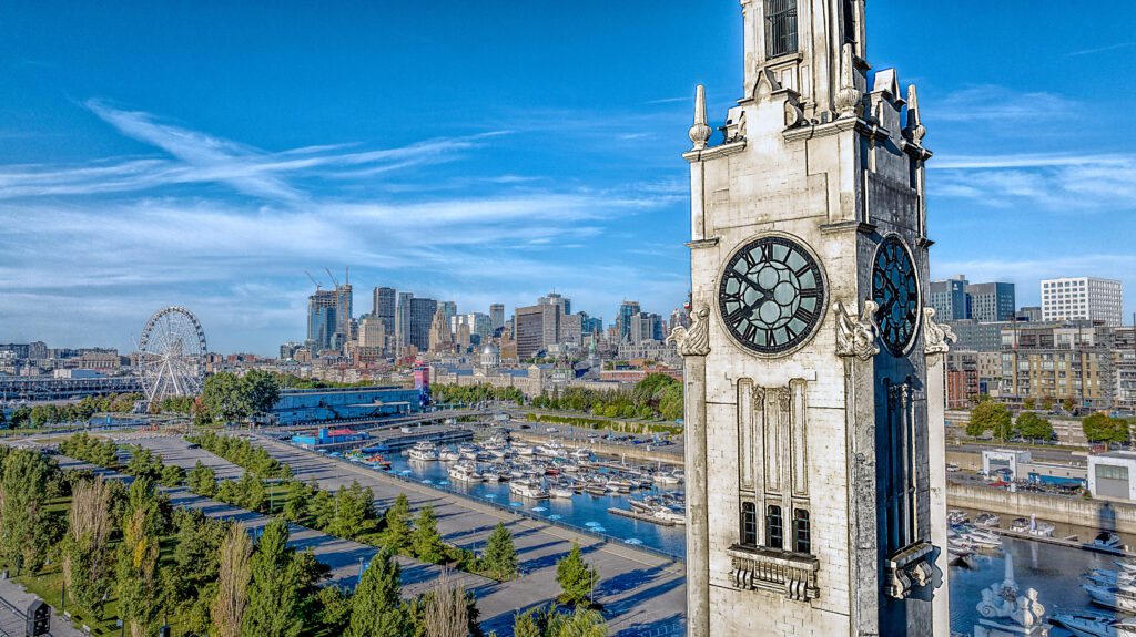 Aerial view of the Clock Tower in Montreal, Canada, with a large clock face visible. In the background, a Ferris wheel, downtown buildings, and a marina with docked boats line the waterfront under a clear blue sky.