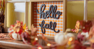 A decorative sign with the words "hello fall" in cursive black letters is set against an orange and white checkered background. The sign is framed in a wooden box and surrounded by autumn-themed decorations, including small white pumpkins and artificial fall leaves. The scene is cozy and festive, capturing the essence of the first day of fall.