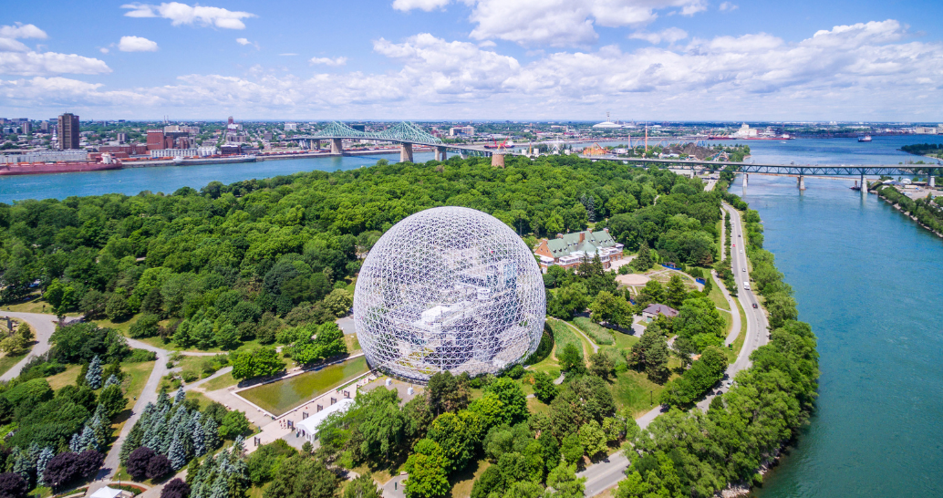 Aerial view of the Montreal Biosphere, a geodesic dome structure surrounded by lush green trees on Saint Helen's Island in Montreal, Canada. The image shows the St. Lawrence River on both sides of the island, with bridges and cityscape visible in the background under a partly cloudy sky.